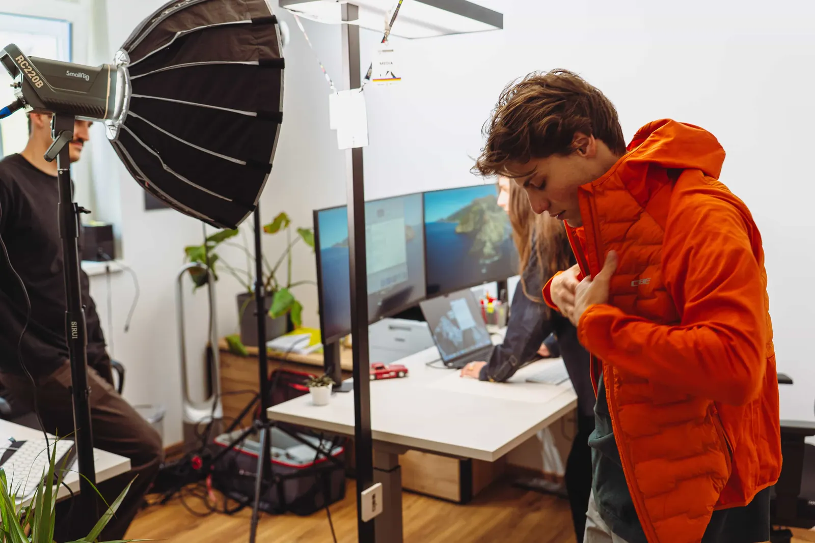 Produktionsaufbau im Innsbrucker Büro von Invisions: Softbox und Dauerlicht vor den Schreibtischen, ein Model zieht eine orange Outdoorjacke an.