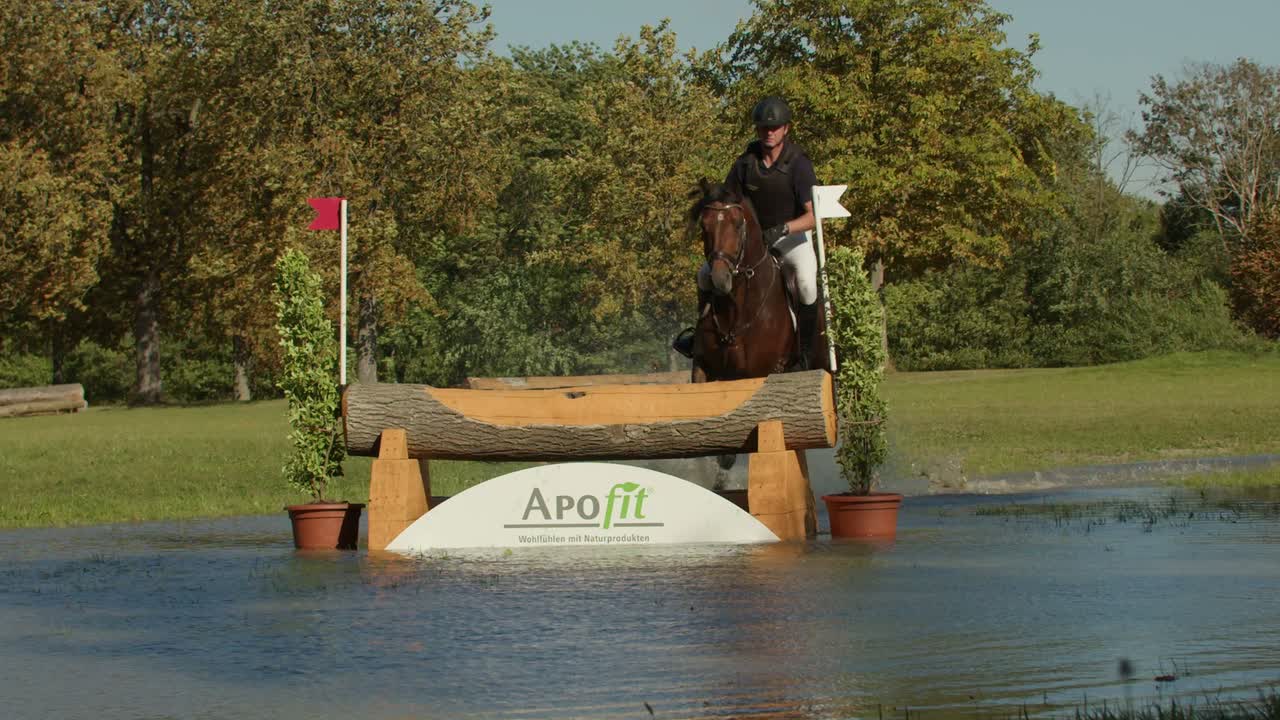 Standbild aus dem Invisions Showreel: ein Pferd springt bei einem Geländeritt über einen Baumstamm ins Wasser, die weiße Bande darunter trägt das Logo von APOfit.
